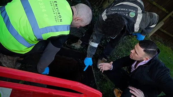 Engineers from Environ Drainage Services examining a drainage inspection chamber, discussing potential issues, with safety gear and a red barrier in the foreground.