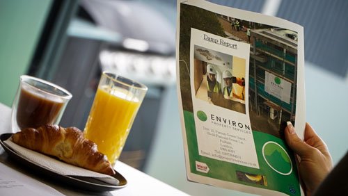 Hand holding an Environ Drainage Services report featuring a drainage inspection image, with a croissant and drinks on a table, emphasizing professional drainage solutions in London.
