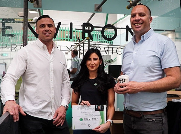 Team members of Environ Property Services posing in their office, showcasing a certificate of achievement, with the company logo prominently displayed in the background.