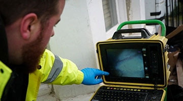 Technician using a CCTV drain inspection camera, pointing at the monitor displaying drainage system visuals, emphasizing Environ Drainage Services' expertise in emergency drain services for stadiums and event spaces.