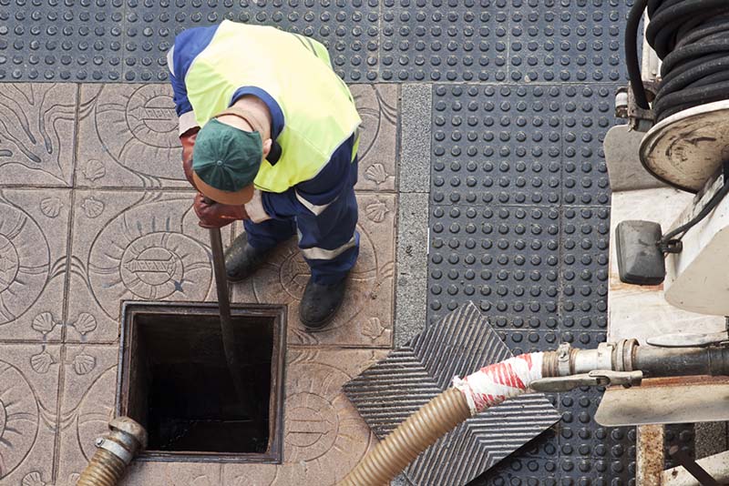 Worker in safety gear performing drain maintenance, using a tool to inspect a drainage system, highlighting the importance of regular drain surveys.