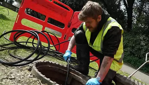 Man in a high-visibility vest using a tool to inspect a drainage system, with drainage cables and safety barriers in the background, illustrating emergency drain services for stadiums.