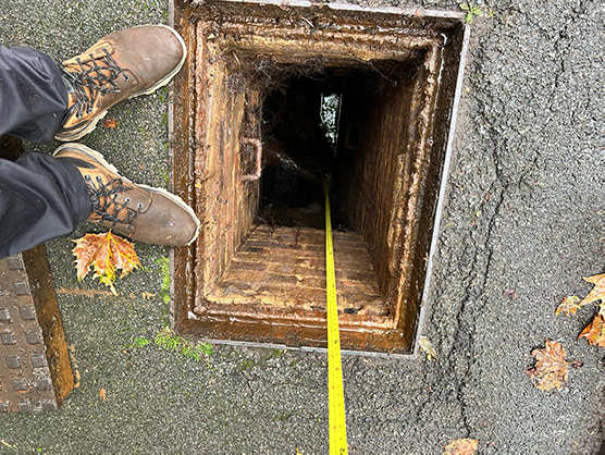 Person standing above a drainage manhole with a measuring tape inside, showcasing commercial drainage inspection and maintenance services.