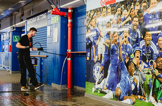 Technician using high-pressure water jet to clear drain blockage in stadium corridor, with vibrant Chelsea FC mural in background, emphasizing professional drain unblocking services for event venues.