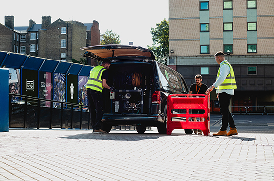 Technicians in high-visibility vests working on drainage solutions at a stadium, with equipment visible in the open van and safety barriers in the foreground.