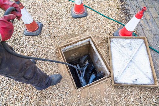 Drainage technician using equipment to inspect a drainage system, with safety cones and a manhole cover in a gravel area, highlighting professional drain services.