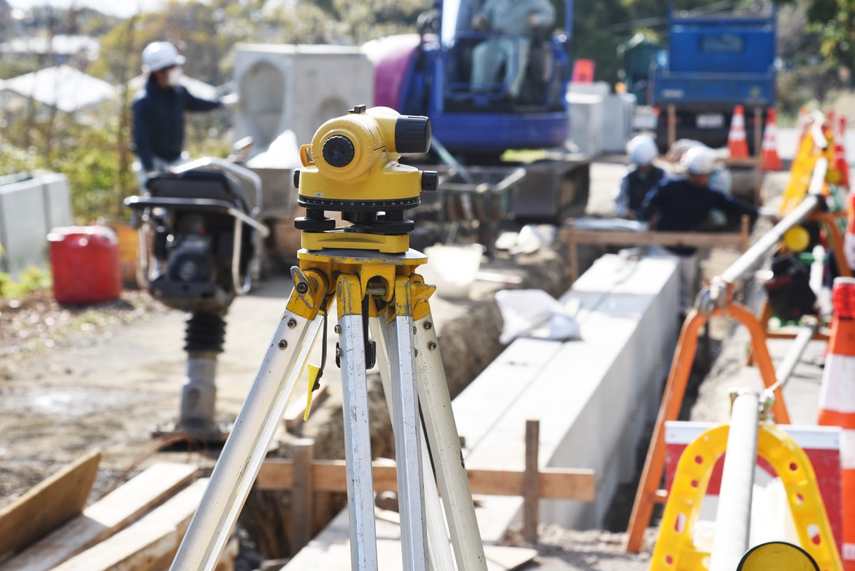 Construction site with a yellow surveying instrument in the foreground, drainage work in progress, workers in safety gear, and construction equipment visible in the background, illustrating professional drainage services.